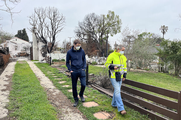 Two people walk down a driveway, a burnt home behind them and grass and trees in early bloom to the side.